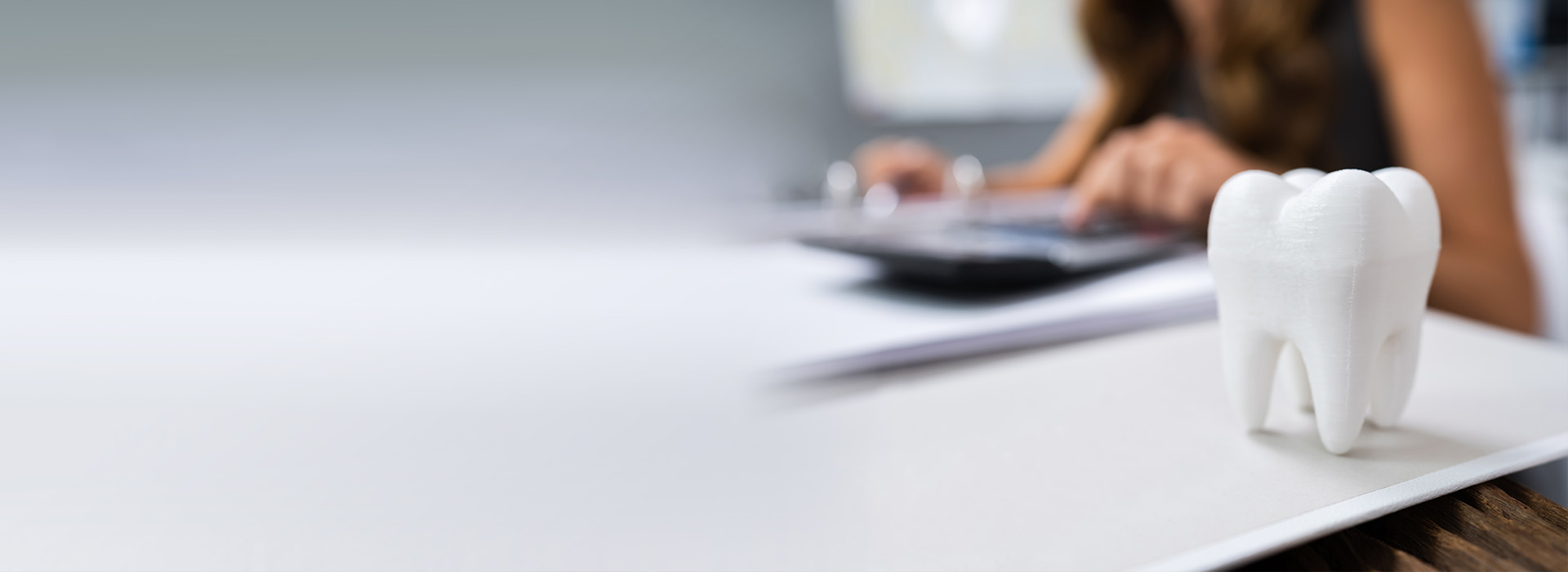 The image is a photograph of a person sitting at a desk with a laptop, papers, and a toothbrush holder in the foreground.