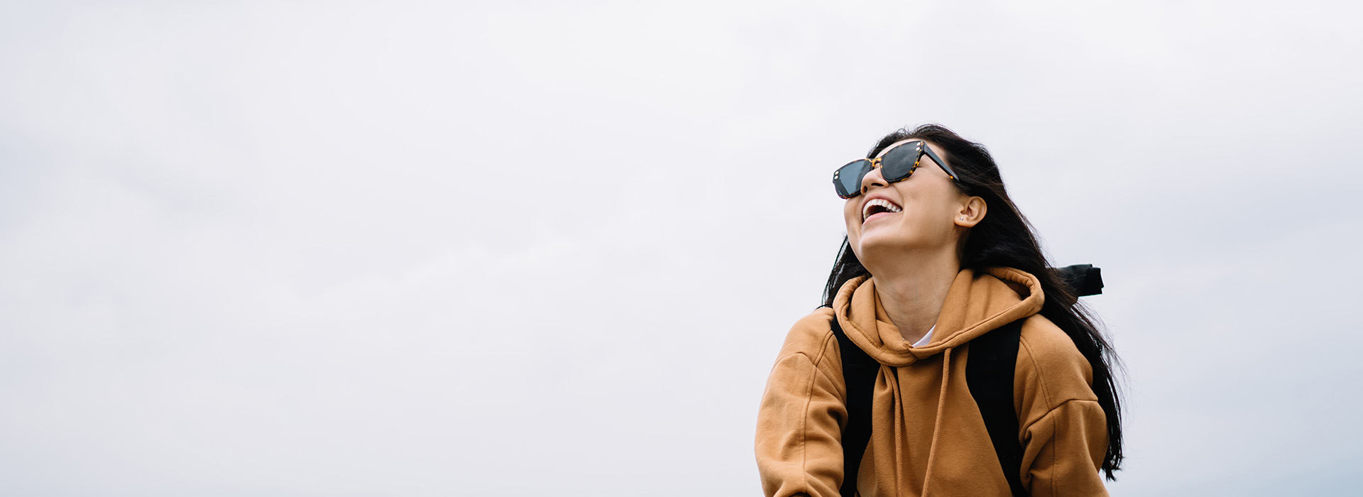 A person in a hoodie and sunglasses stands against a cloudy sky, smiling.