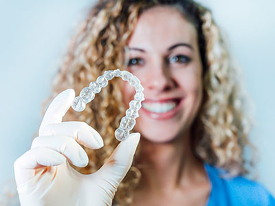 A woman in a white lab coat is holding up a dental retainer with a smile, showcasing its transparent nature.