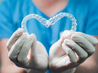 The image shows a pair of clear braces being held by hands wearing white gloves, with the background featuring a blue medical gown and a heart-shaped object that resembles a piece of dental equipment.