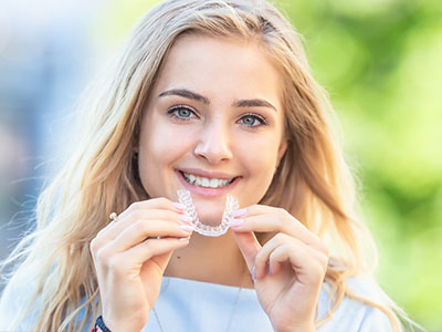 A young woman is holding a clear dental retainer, smiling and looking directly at the camera.