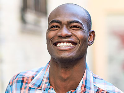 The image features a man with a radiant smile, wearing a blue and white plaid shirt, against a backdrop of a building facade.