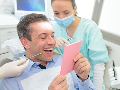 The image depicts a man holding a pink card while sitting in an office chair, with a dental professional wearing gloves and a surgical mask behind him.