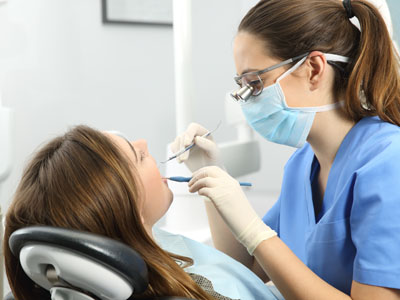 A dental hygienist is performing a cleaning procedure on a seated patient in a dental office, with both wearing protective gear.