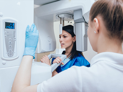 A woman in a blue coat and white gloves inspects a large, modern CT scanner with a digital display, while another person stands behind it.