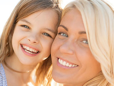 A smiling woman and a young girl, both with blonde hair, sharing a joyful moment together against a sunlit backdrop.