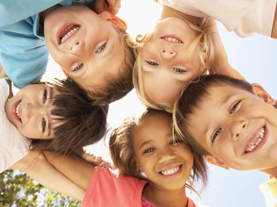 The image shows a group of children, likely in their early to late childhood years, smiling and posing together for a photo against a clear sky background.