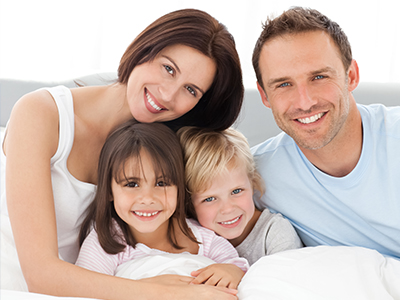 The image shows a family of four, including two adults and two children, smiling together in bed.