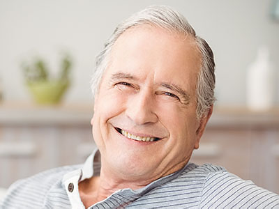 The image shows an elderly man with a smile, sitting in a chair indoors.