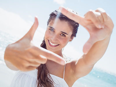 A woman with long hair, wearing a white top, is taking a selfie with her right hand against a sunny beach backdrop.