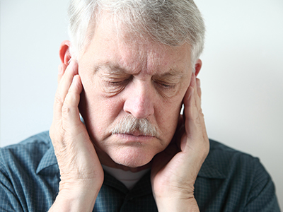 An elderly man with a mustache, wearing glasses and a blue shirt, holding his head in pain or discomfort.